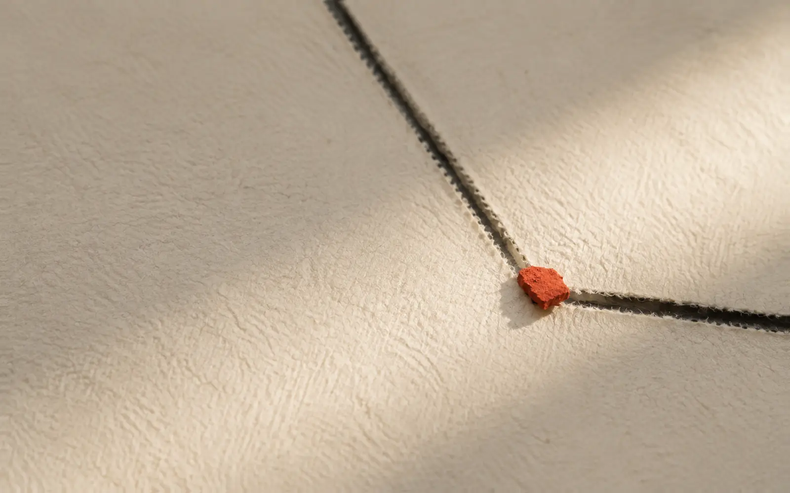 Two stitched seams on bone paper meet at one small red fragment in raking light.