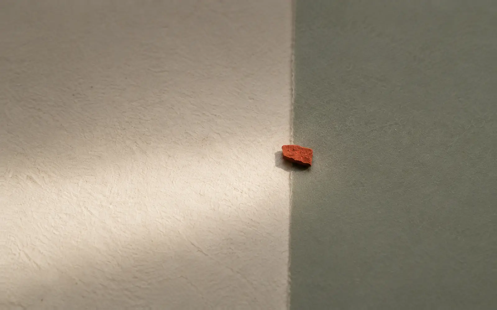 A sheet of bone paper meets a sheet of sage paper at a fold; a small red fragment rests on the fold in raking light.